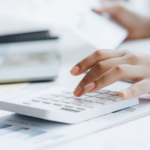 Hände tippen auf einen Taschenrechner, im Hintergrund ein Laptop und Unterlagen auf einem Tisch