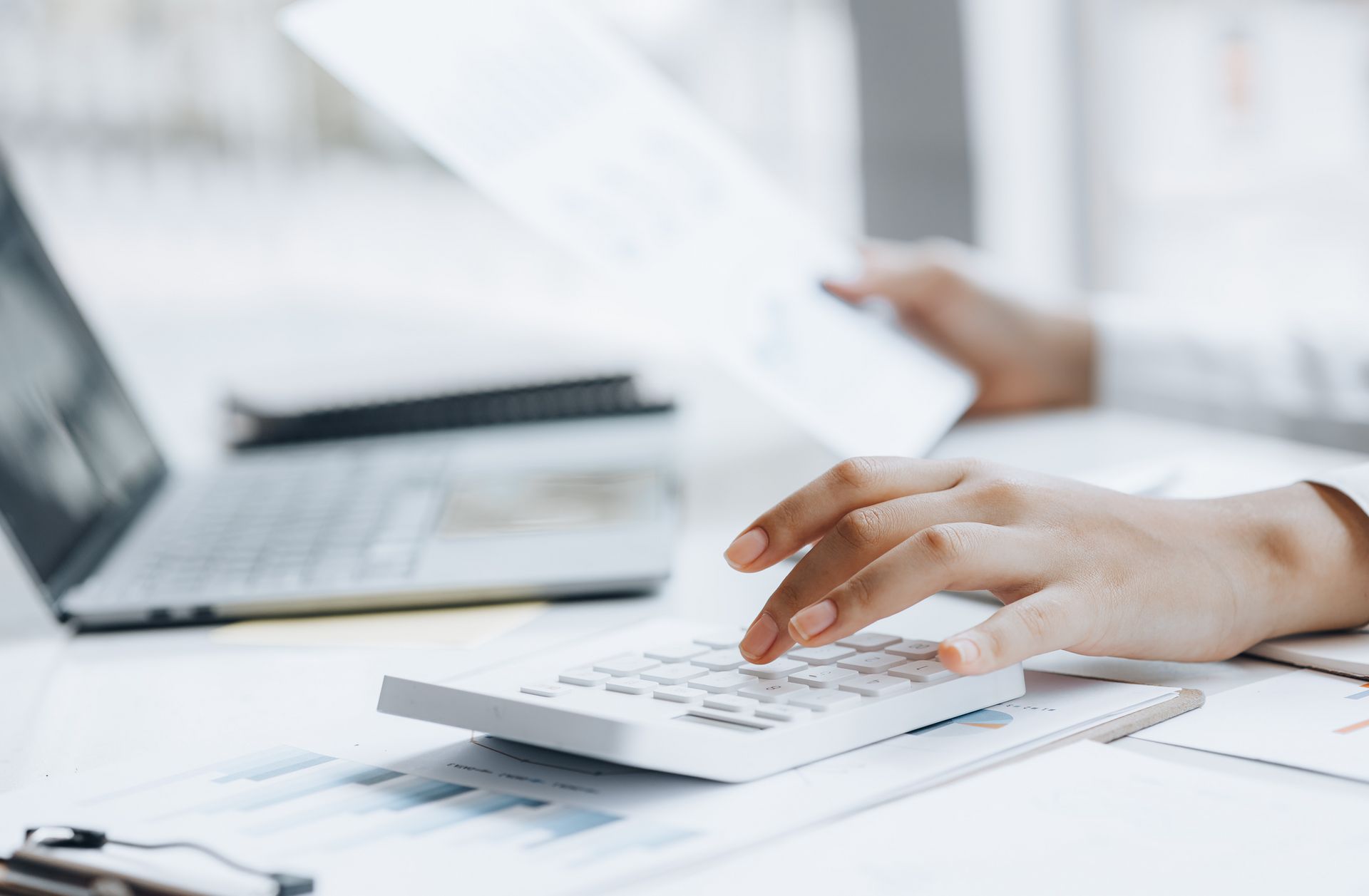 Hände tippen auf einen Taschenrechner, im Hintergrund ein Laptop und Unterlagen auf einem Tisch