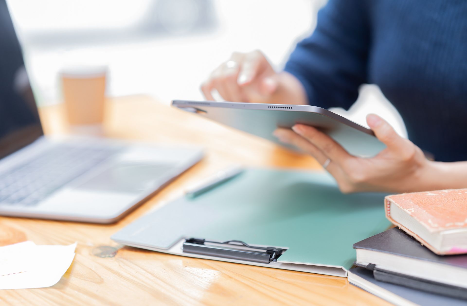 Person hält Tablet über Schreibtisch mit Laptop, Klemmbrett und Büchern