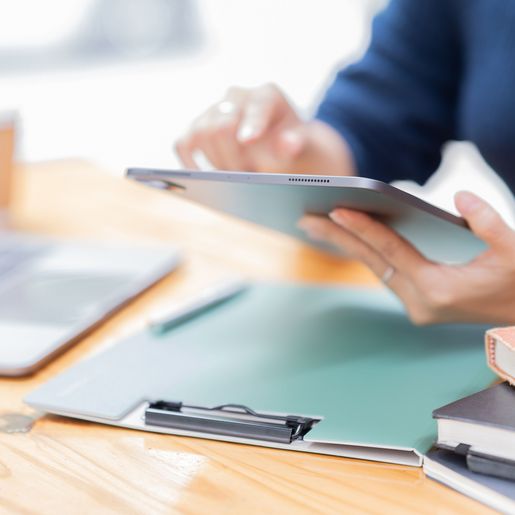 Person hält Tablet über Schreibtisch mit Laptop, Klemmbrett und Büchern
