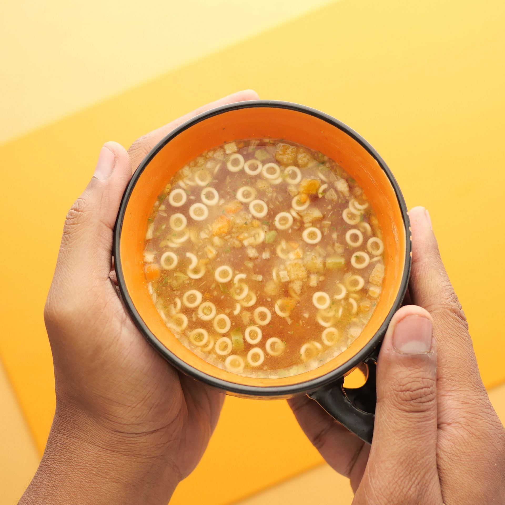 Two hands holding an orange bowl of dry instant noodles in the shape of letters on a yellow background