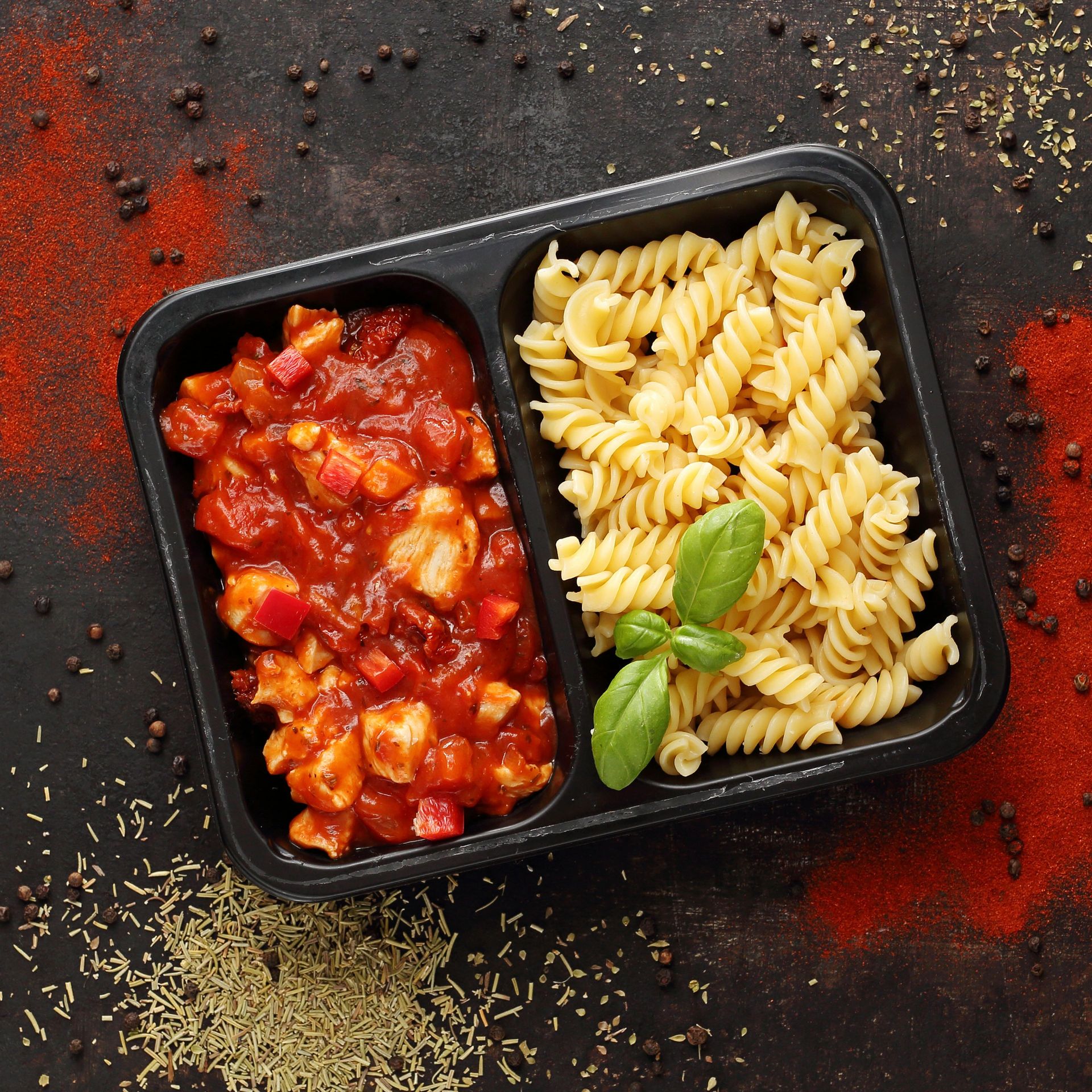 Black plastic bowl with pasta and tomato sauce, garnished with basil, surrounded by powdered spices