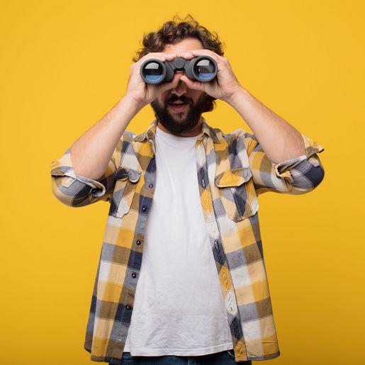 Mann mit Bart und lockigem Haar schaut durch Fernglas vor gelbem Hintergrund